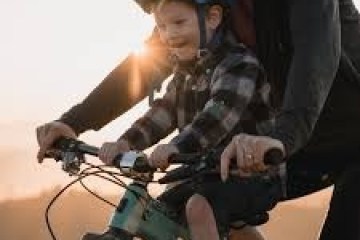 Child wearing a helmet rides a bike with an adult in warm sunlight.