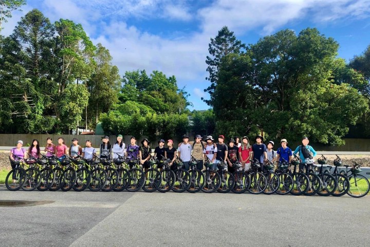 Group photo of people with bicycles lined up on a road with trees in the background.