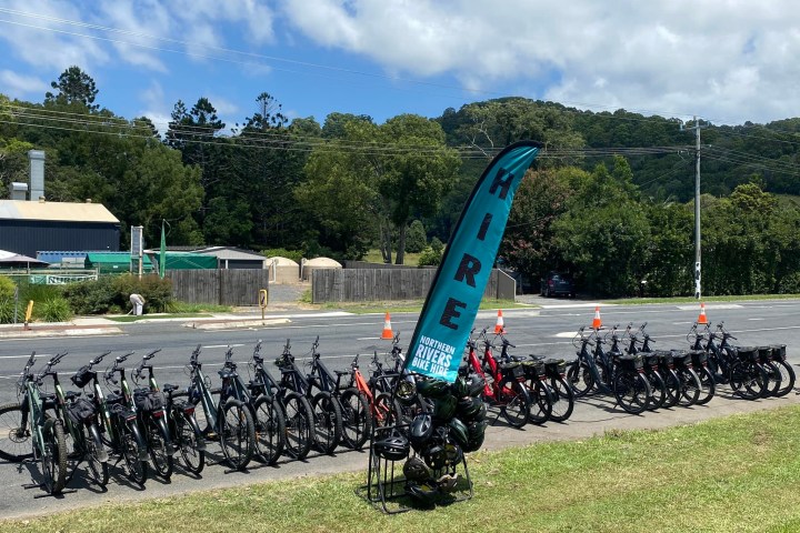 Row of bicycles with a 'Hire' sign by a road, surrounded by trees and a clear sky.