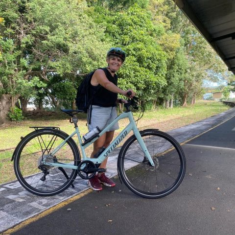 Person smiling with helmet standing beside a bike on a pathway with trees in the background.