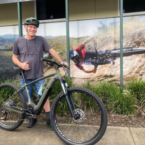 Man with helmet posing next to mountain bike in front of cycling-themed mural.