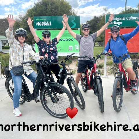 Four people on bikes raising arms in front of Mooball sign.