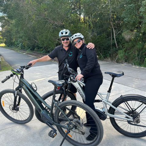 Two cyclists wearing helmets stand with their bikes on a paved path.