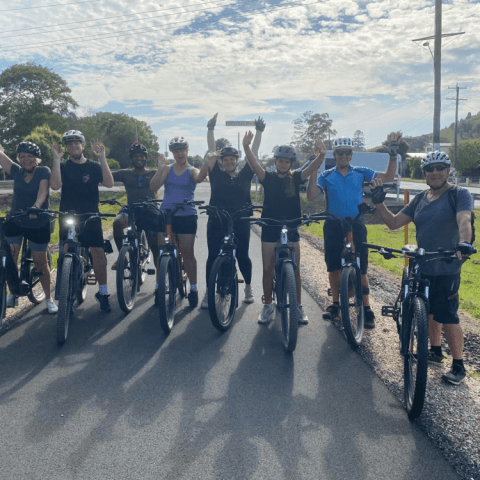 Group of cyclists posing on a road with bicycles, arms raised, under a partly cloudy sky.