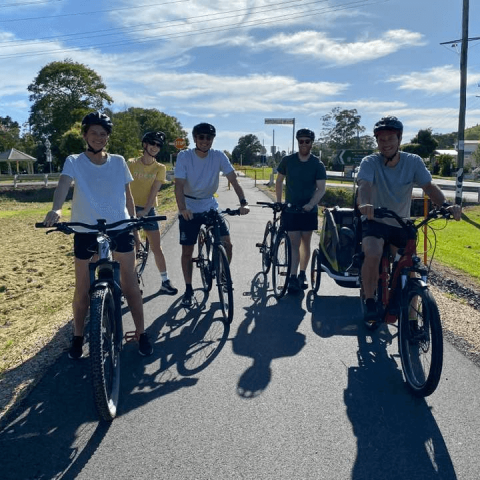 Five people on bicycles with helmets, smiling on a sunny pathway.