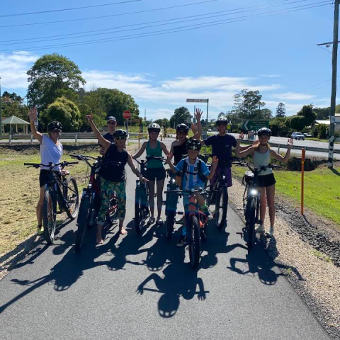 Group of eight cyclists on a path, waving at the camera, with trees and road signs in the background.