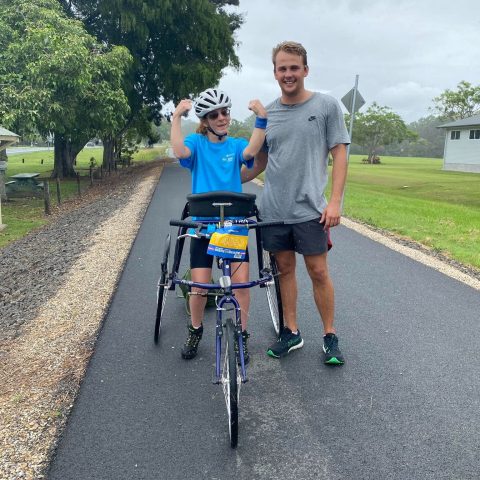 Two people on a road, one using a tricycle, both smiling and flexing muscles.
