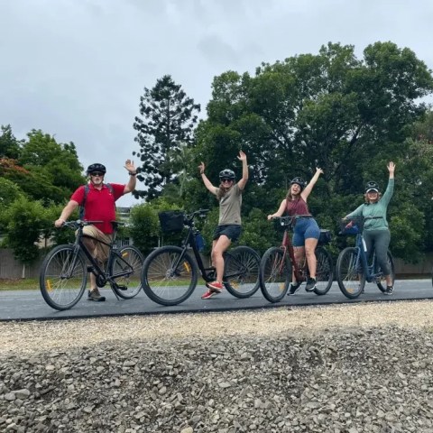 Four people on bicycles, wearing helmets, raising hands, with trees in the background.