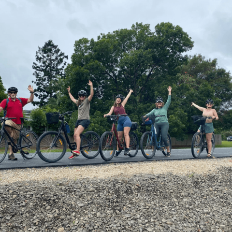 Five people on bikes raising hands, trees in background, overcast sky.