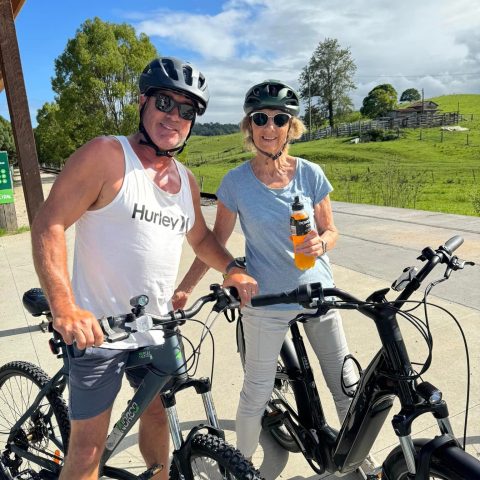 Two people wearing helmets stand with bicycles, smiling on a sunny day with a grassy field in the background.