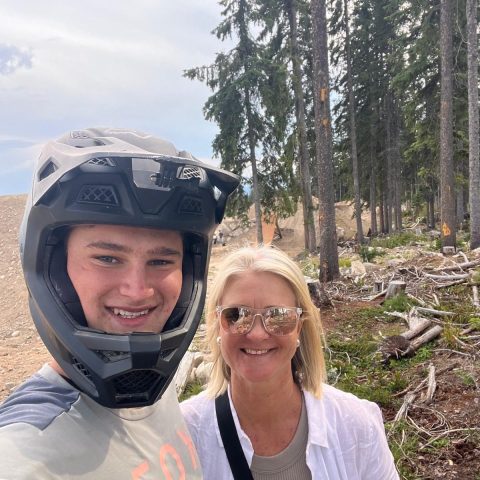 Two people outdoors, one in a helmet, both smiling in a forested area with fallen logs.