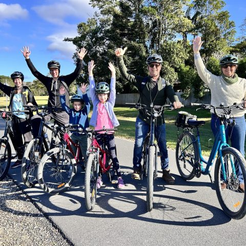 Group of six people with bicycles raising arms on a sunny day.