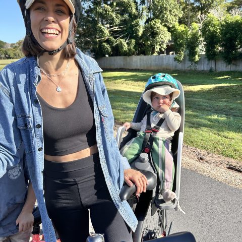 Woman smiling with toddler in bike seat, both wearing helmets, outdoor setting.