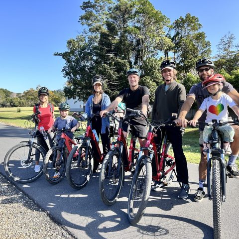 Group of seven people with bicycles, wearing helmets, standing on a paved path with trees in the background.