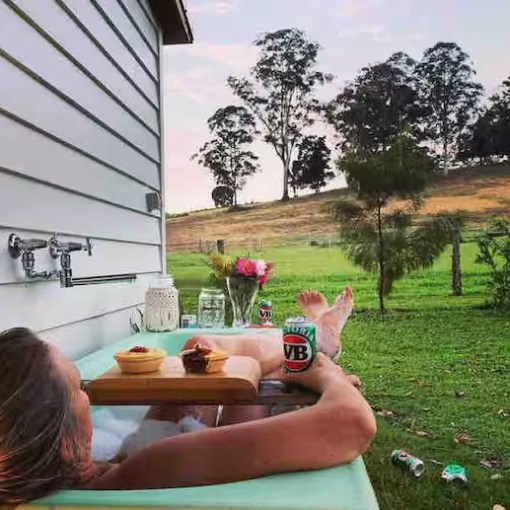 Person relaxing in an outdoor bathtub with drinks and snacks, overlooking a grassy field and trees.
