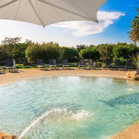 Poolside view with sun loungers, umbrella, and trees under a clear blue sky.