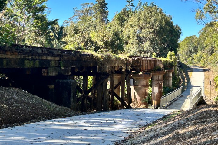 Old wooden bridge over a forested path with trees and clear sky.