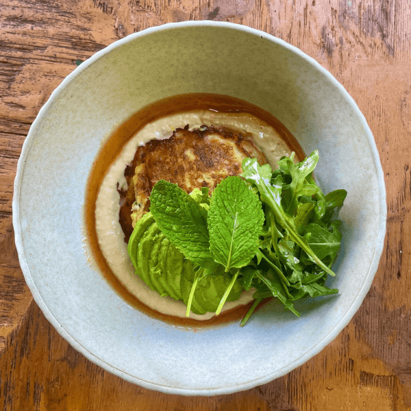 A bowl with a savory dish featuring a patty, avocado, arugula, and dressing on a wooden table.