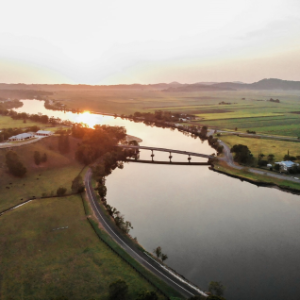 Aerial view of a river with a bridge at sunset, surrounded by fields and a few buildings.