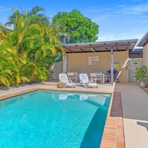 Outdoor pool with lounge chairs, tropical plants, and a covered seating area under a sunny sky.
