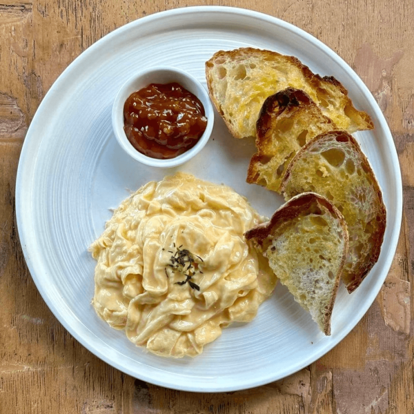 Plate with creamy scrambled eggs, sliced toasted bread, and a cup of chutney on a wooden table.