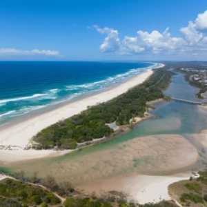 Aerial view of a beach with a river, sand dunes, and greenery under a partly cloudy sky.