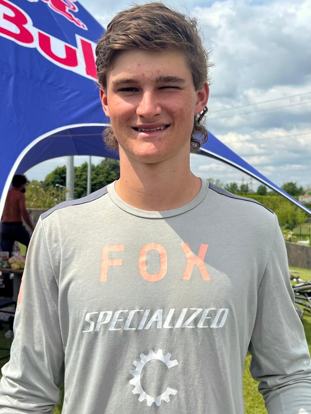 Person in a gray shirt with 'FOX' and 'SPECIALIZED' logos, standing outdoors by a Red Bull tent.
