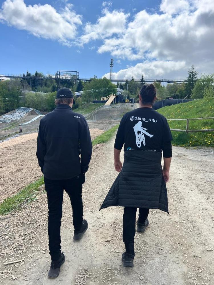 Two men walking on a dirt path at a biking park under a partly cloudy sky.