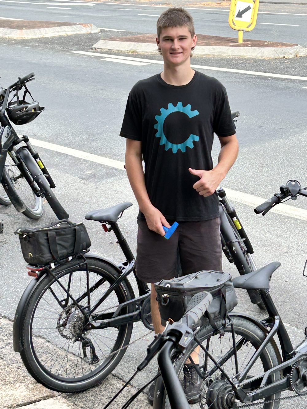 Person smiling next to bicycles on a street, wearing a black shirt with a blue gear logo.