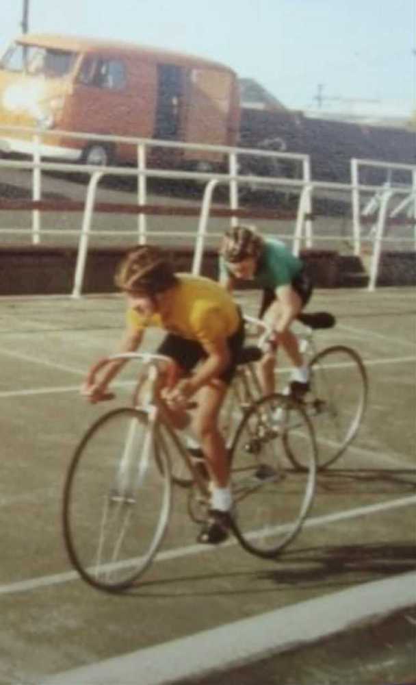 Two cyclists racing on a track, yellow jersey in front, with a parked van in the background.