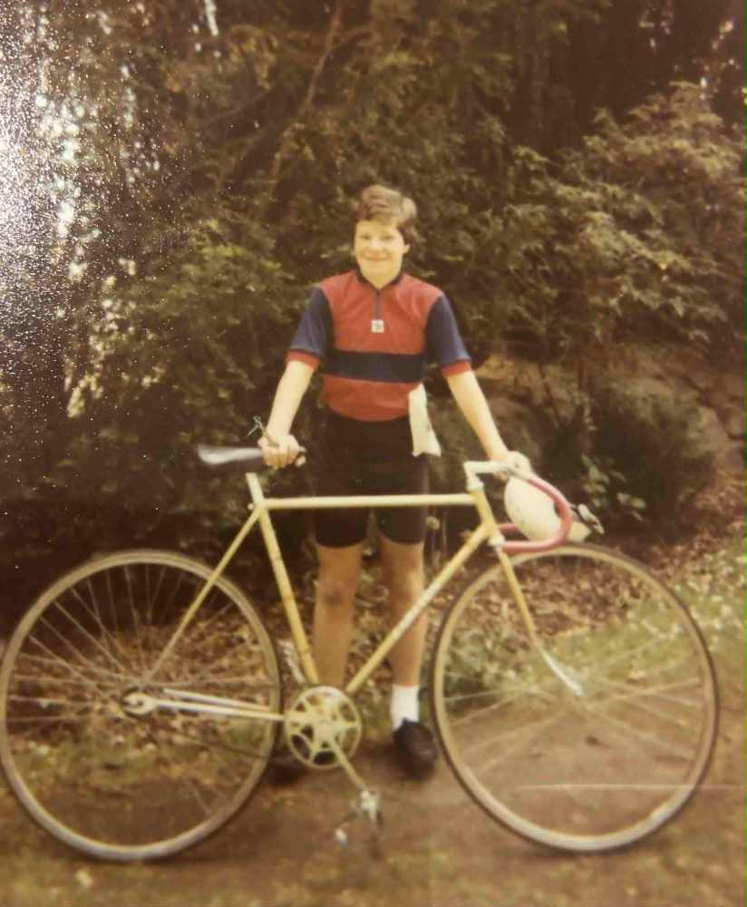 Person in cycling outfit stands with vintage bike in a garden.