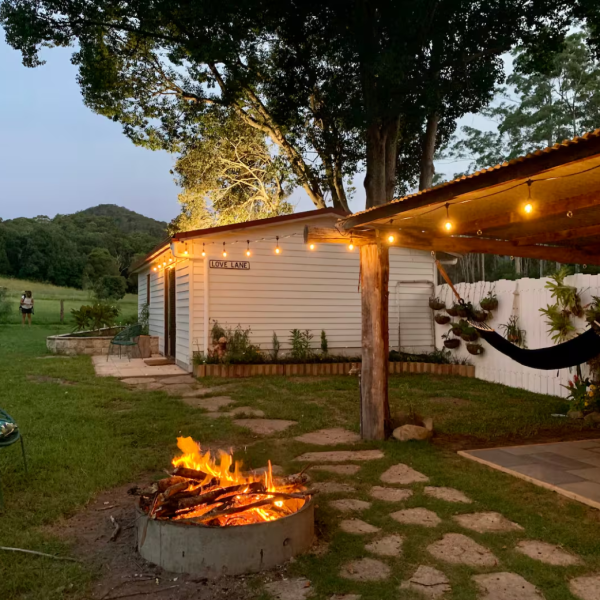 Outdoor scene with a fire pit, hammock, string lights, trees, and a small cabin at dusk.