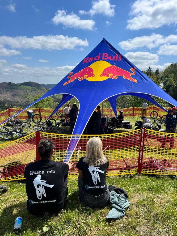 Two people sitting near Red Bull tent, bikes, and blue sky.