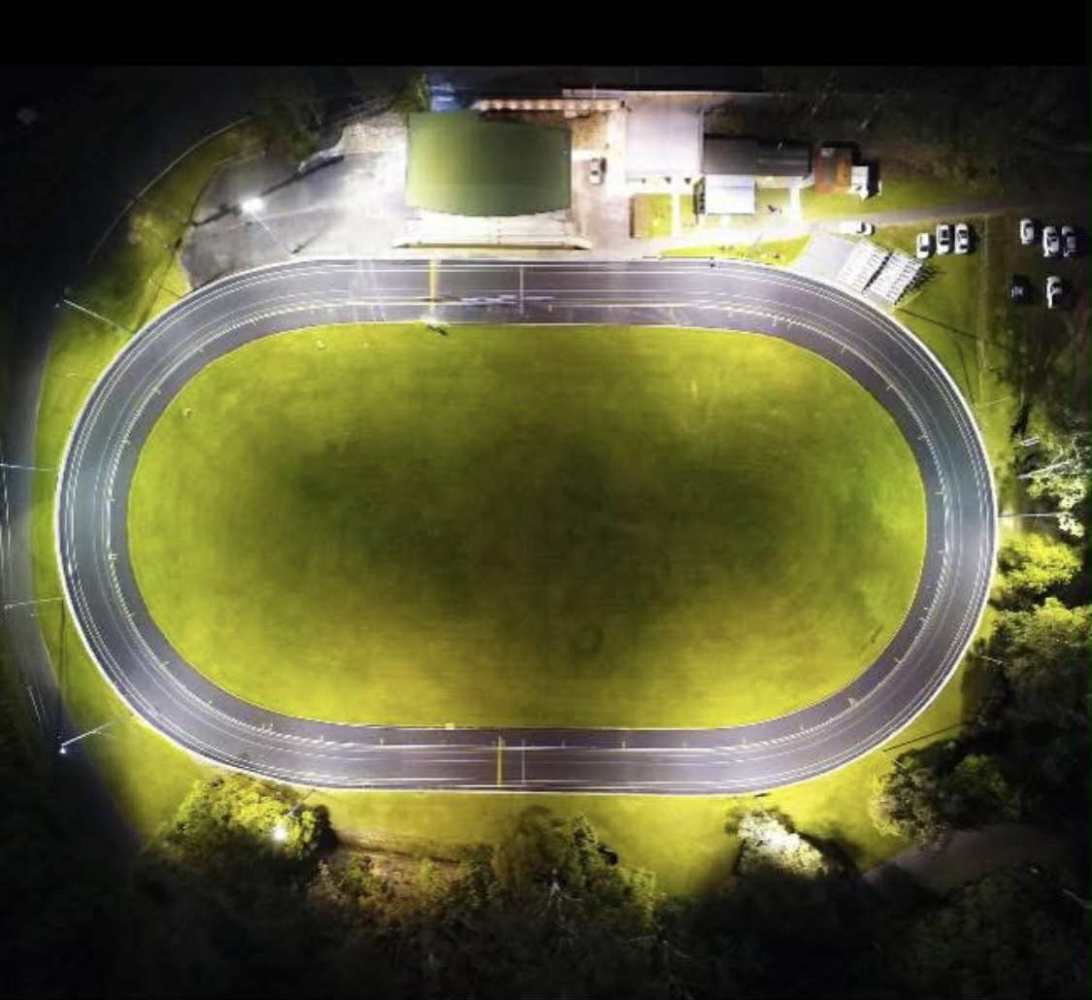 Aerial view of an illuminated asphalt track oval on grass, at night.