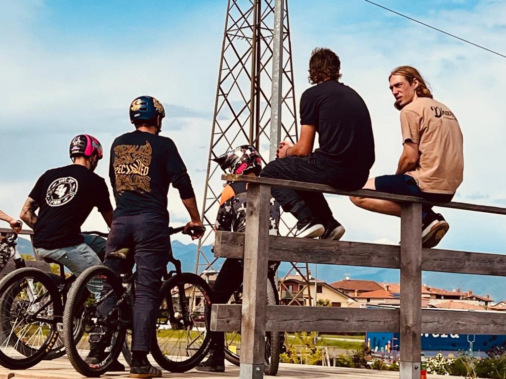 Three cyclists and two seated men chatting by a wooden railing under a power line.