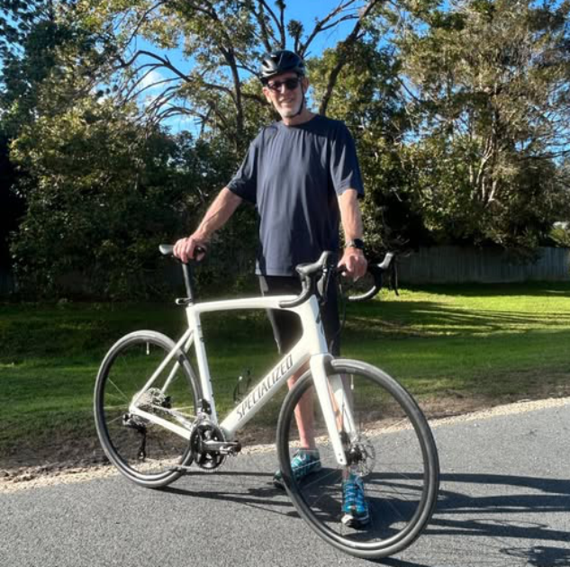 Person with helmet poses holding a white bicycle on a path with trees in the background.